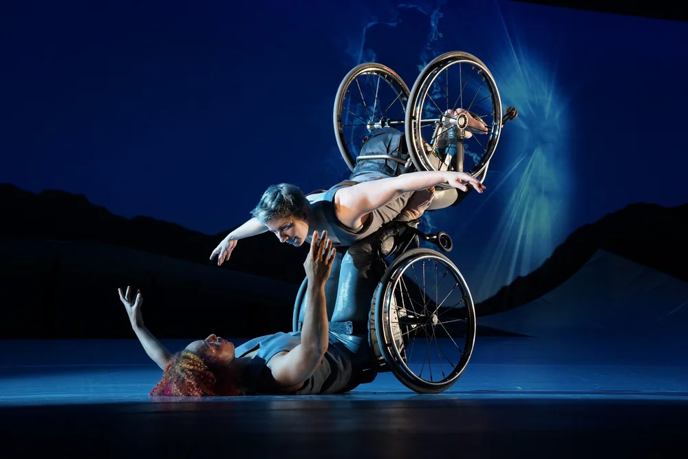 Image Alt text: Laurel Lawson as Venus is flying in the air with arms spread wide, wheels spinning, and supported by Alice Sheppard as Andromeda who is lifting from the ground below. They are making eye contact and smiling. Photo by Jay Newman / BRITT Festival.