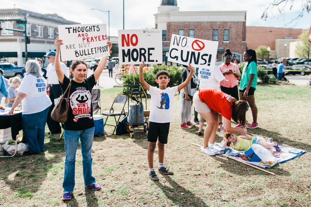 Photo by Heather Mount on Unsplash . A North Texas mother and her son hold protest signs at the March for Our Lives sister rally in Denton, Texas on March 24th, 2018. In the background, volunteers register people to vote.