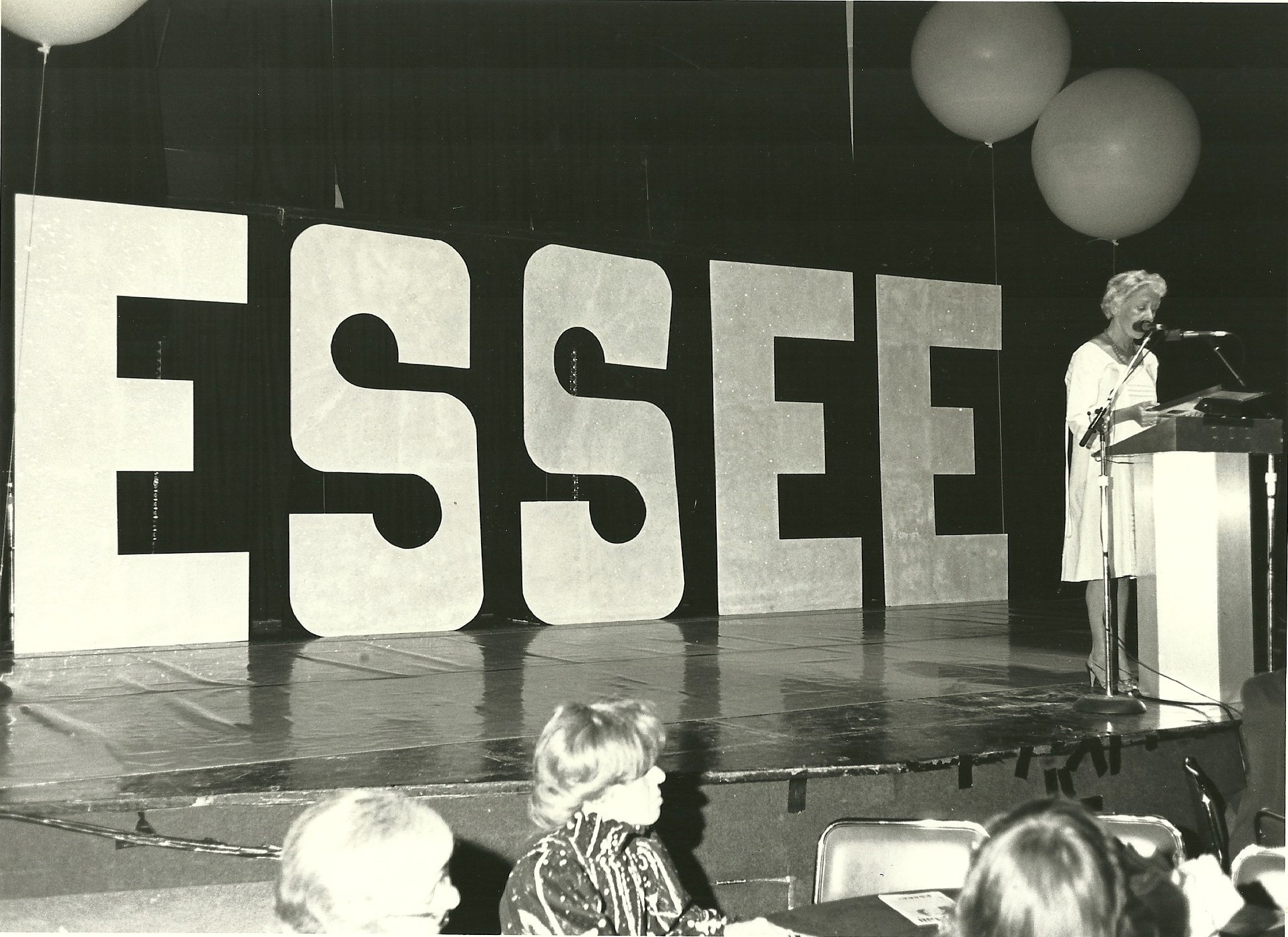 A stage with large block letters spelling out "ESSEE" in the background. The scene is of Mayor Bryne's proclamation for Essee Kupcinet in 1981.