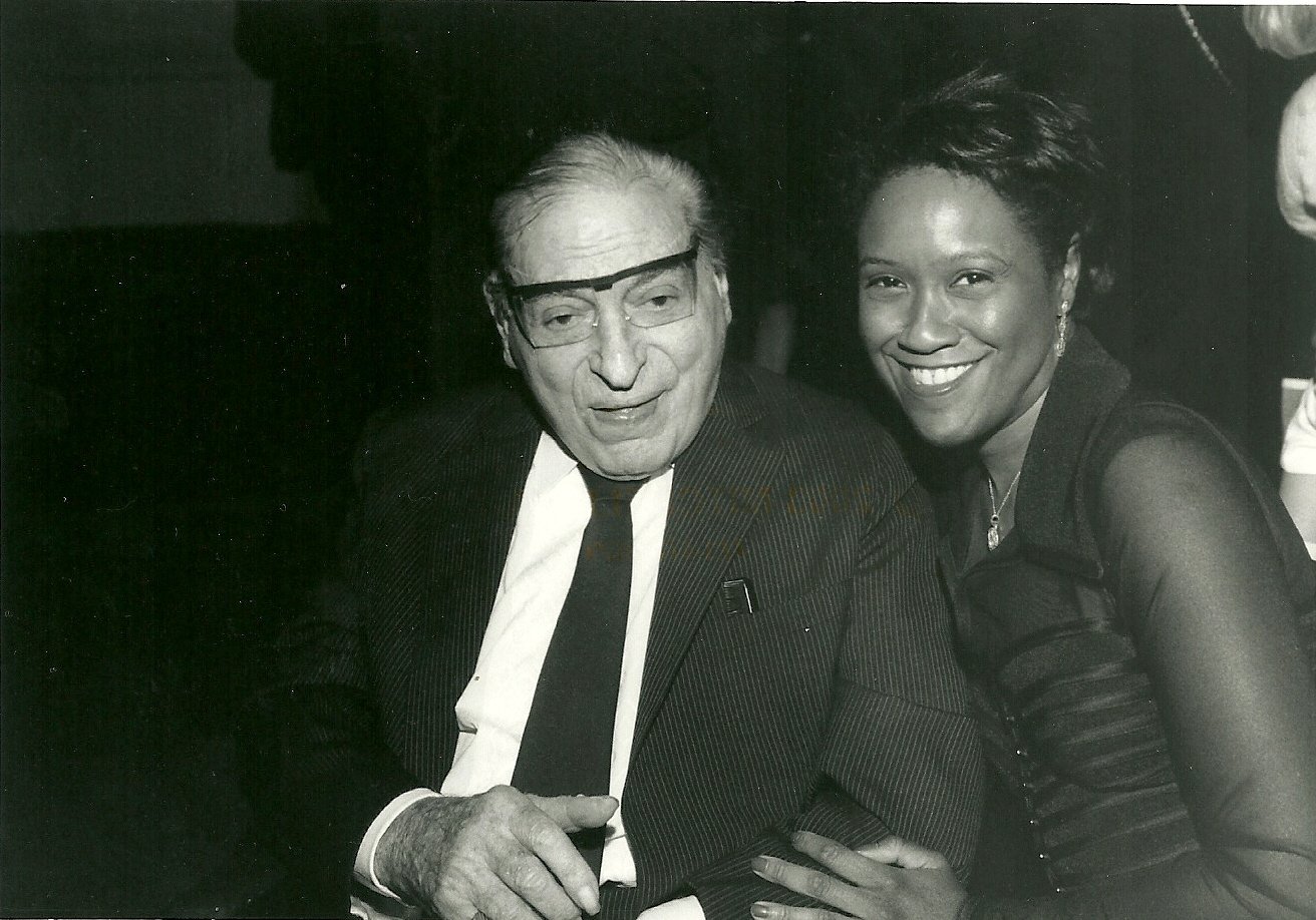 Irv Kupcinet with Pam Jordan at the celebration of Kup’s Column’s 60th anniversary and Irv Kupcinet’s 90th birthday at The Chicago Sun-Times. Irv stands to the left and Pam at the right. They are both smiling at the camera, posed for the picture.