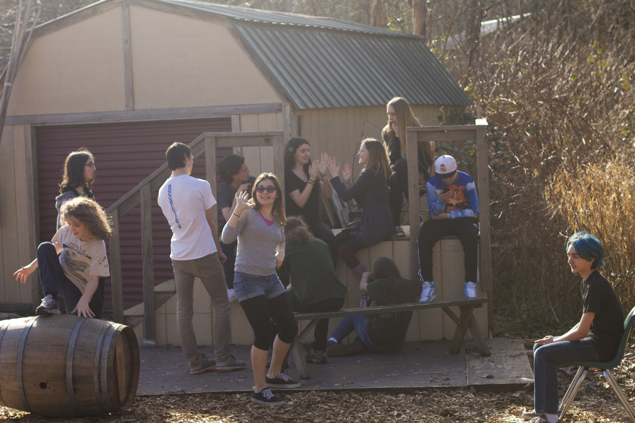Large group of students on an outside structure, crowded around two students who are playing a hand-clapping game.