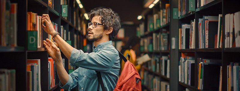 person wearing backpack reaches for book on shelf at library