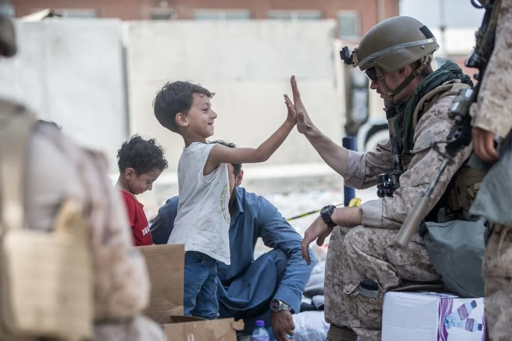 A Marine with Special Purpose Marine Air-Ground Task Force-Crisis Response-Central Command (SPMAGTF-CR-CC) gives a high five to a child during an evacuation at Hamid Karzai International Airport, Kabul, Afghanistan, Aug. 22. U.S. service members are assisting the Department of State with an orderly drawdown of designated personnel in Afghanistan. (U.S. Marine Corps photo by Sgt. Samuel Ruiz).