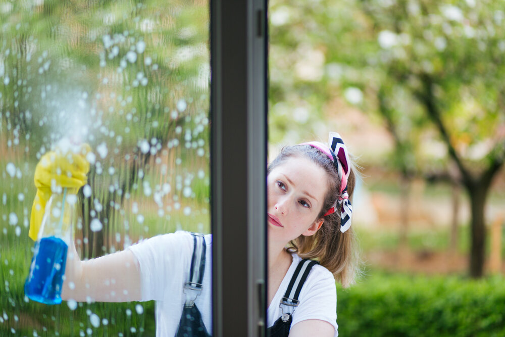 年轻漂亮的女人用喷沫洗洁精瓶子擦窗户. 打扫房子的概念.jpg