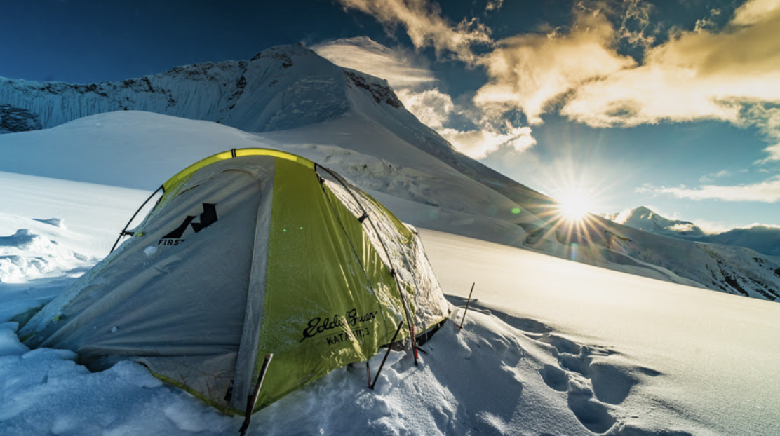 Green tent in the snow with sun setting behind mountains