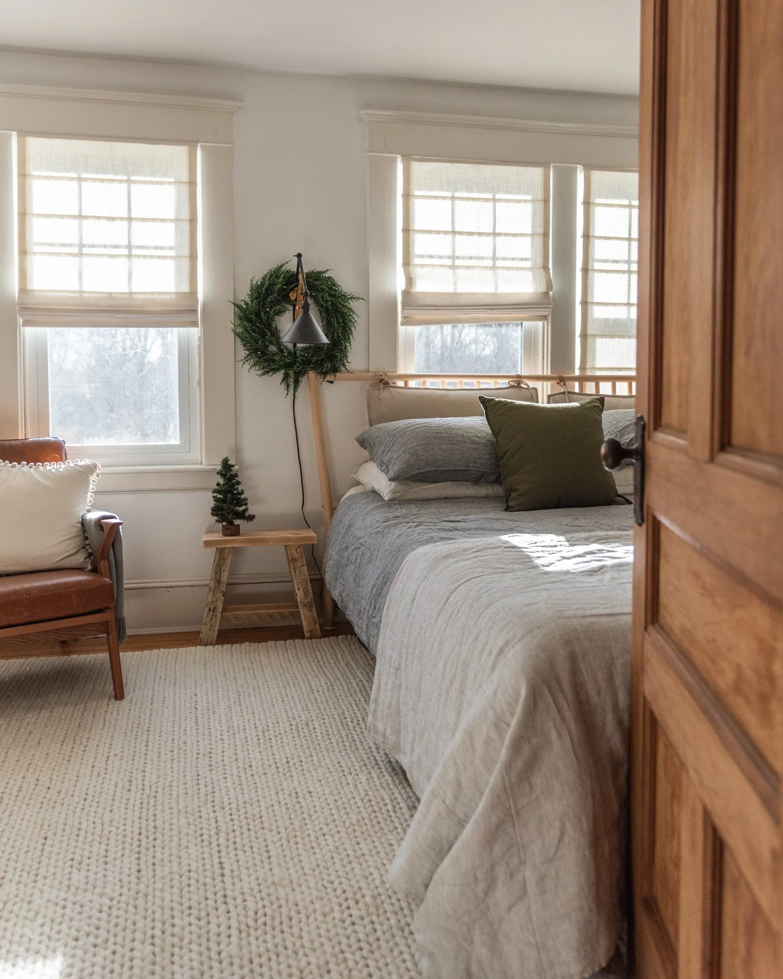 The Wild decoelis | 2020 Holiday Home Tour | Master bedroom with a king size IKEA bedframe in light wood, white linen shades, black sconces with Reese, antique side table milking stool, gray linen bedding. White braided rug USA Leather chair