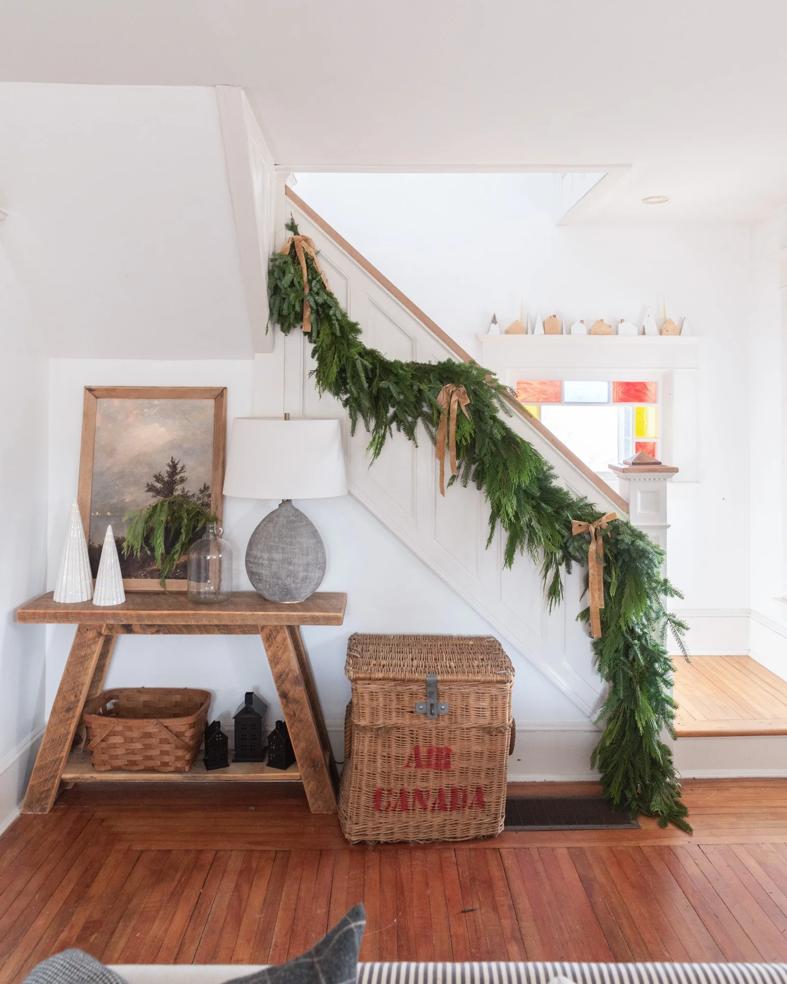 The Wild decoelis | 2020 Holiday Home Tour | Stair garland made out of pine and cedar with gold bows and twinkle lights. White couch with wool throw pillows, vintage wool rug layered over braided wool rug