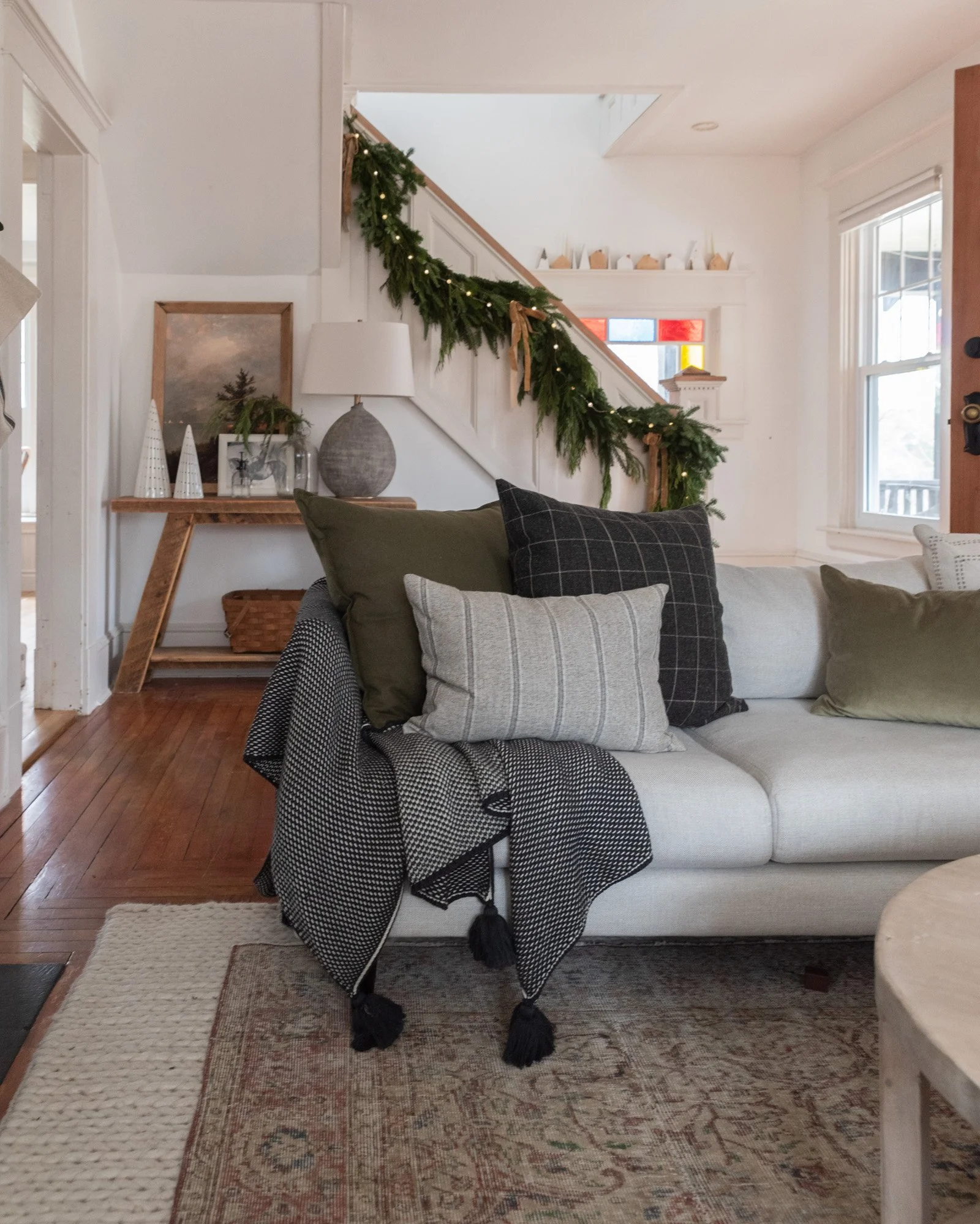 The Wild decoelis | 2020 Holiday Home Tour | Stair garland made out of pine and cedar with gold bows and twinkle lights. White couch with wool throw pillows, vintage wool rug layered over braided wool rug