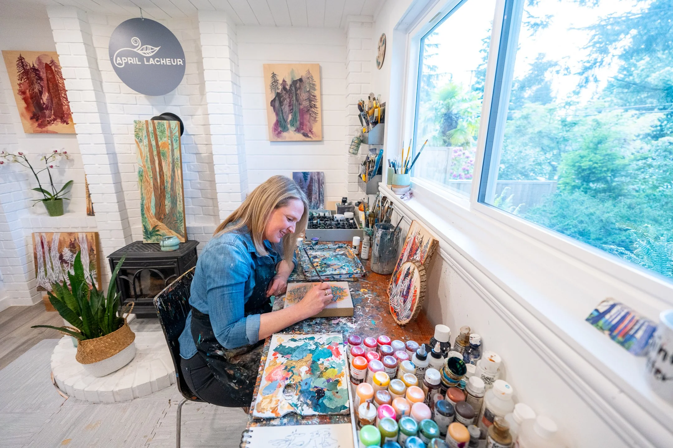 A woman painting in an art studio with paintings on the wall and a large window showing greenery outside.