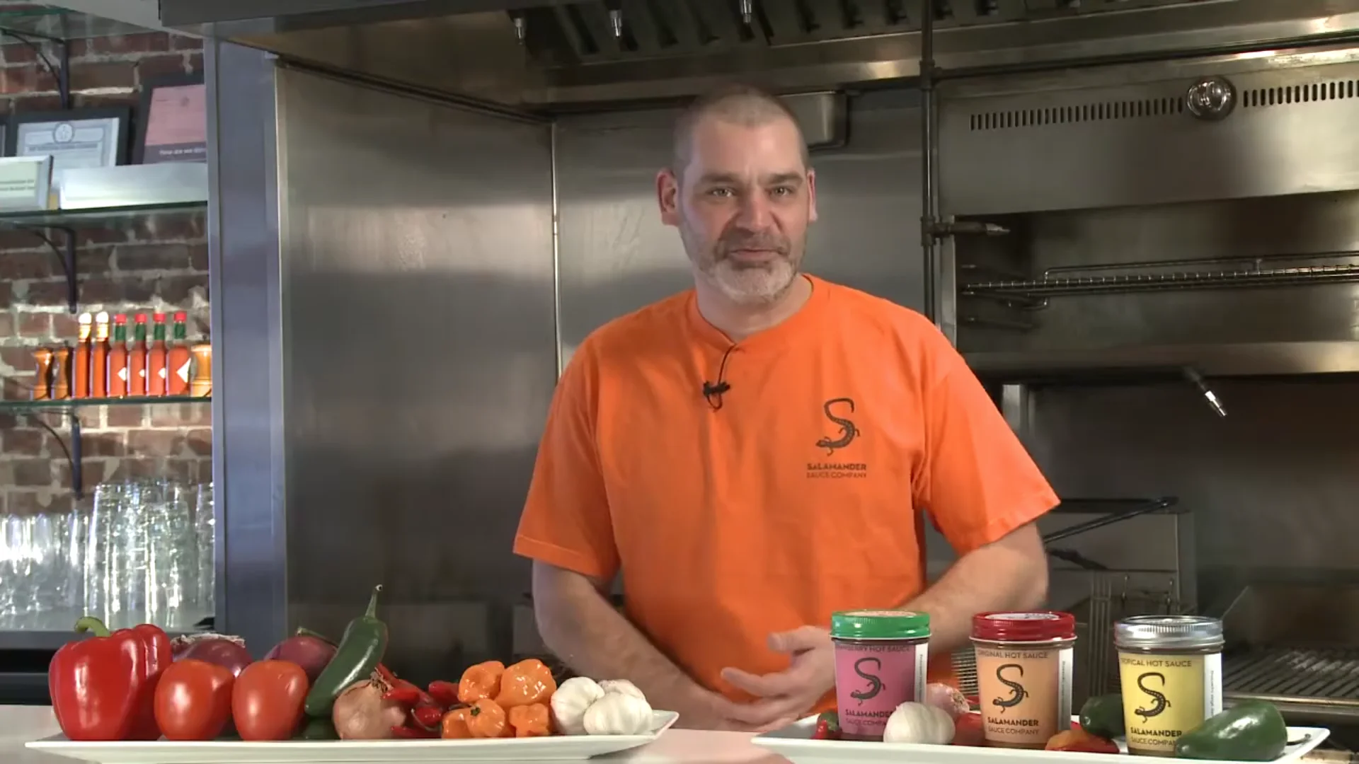 Timothy Kavarnos, founder of Salamander Sauce Company, in a commercial kitchen. Bottles of sauce on the shelf behind him and a salamander broiler over his shoulder.