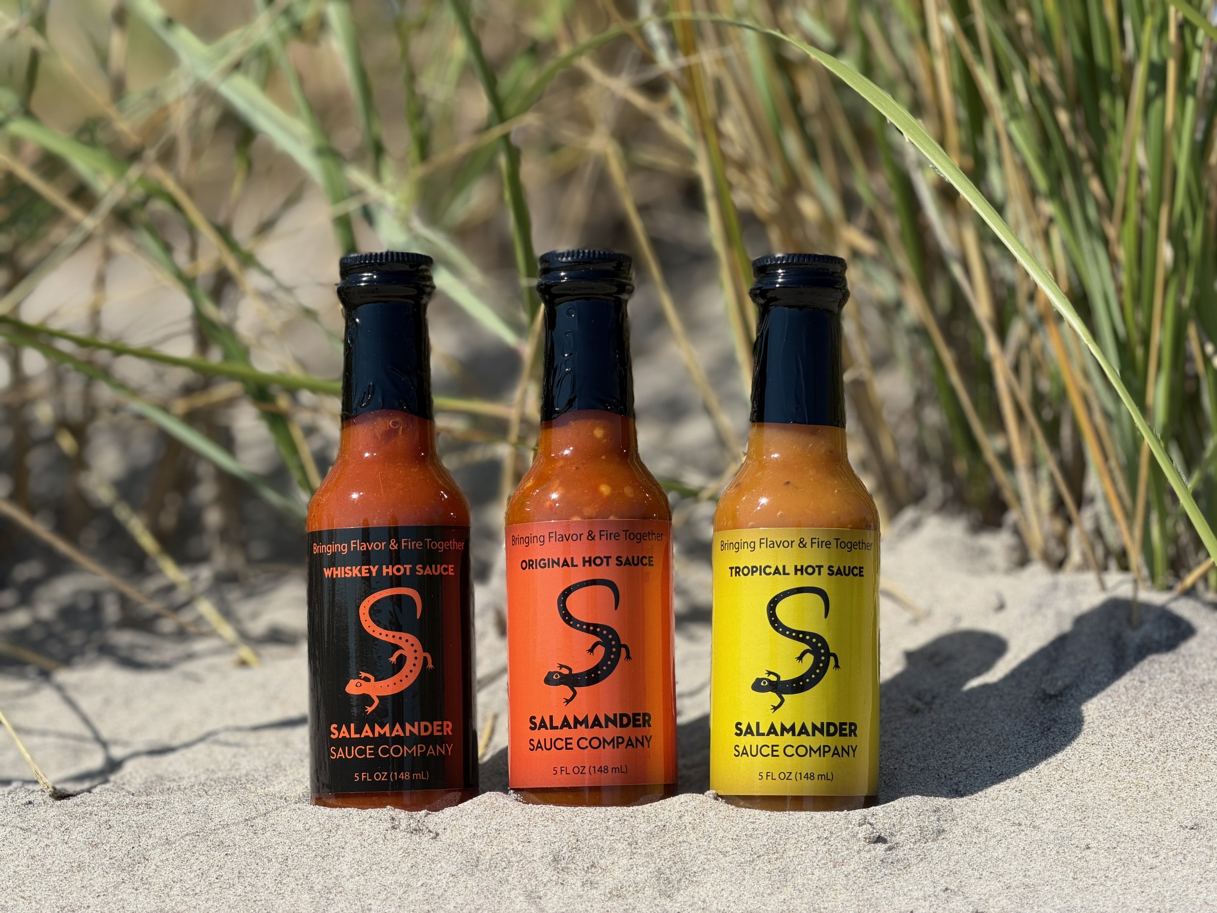 Salamander Sauce Company Whiskey, Original and Tropical hot sauce bottles standing in beach sand in front of dune grass