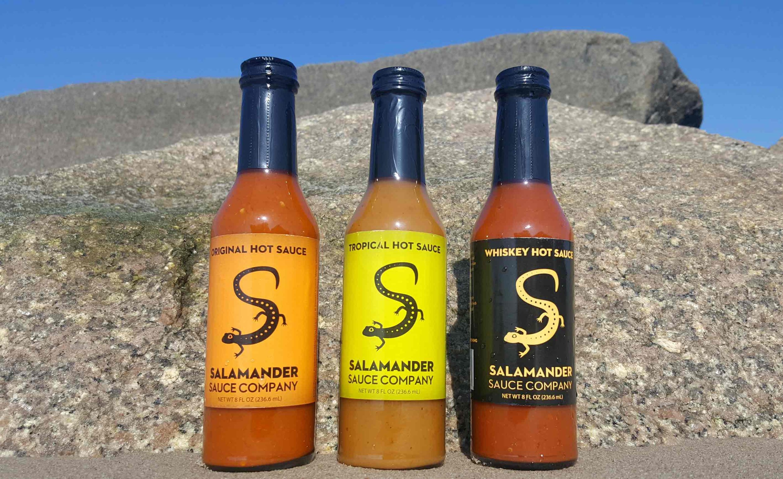 Salamander Original, Tropical and Whiskey hot sauce bottles standing on a granite boulder against an open blue sky