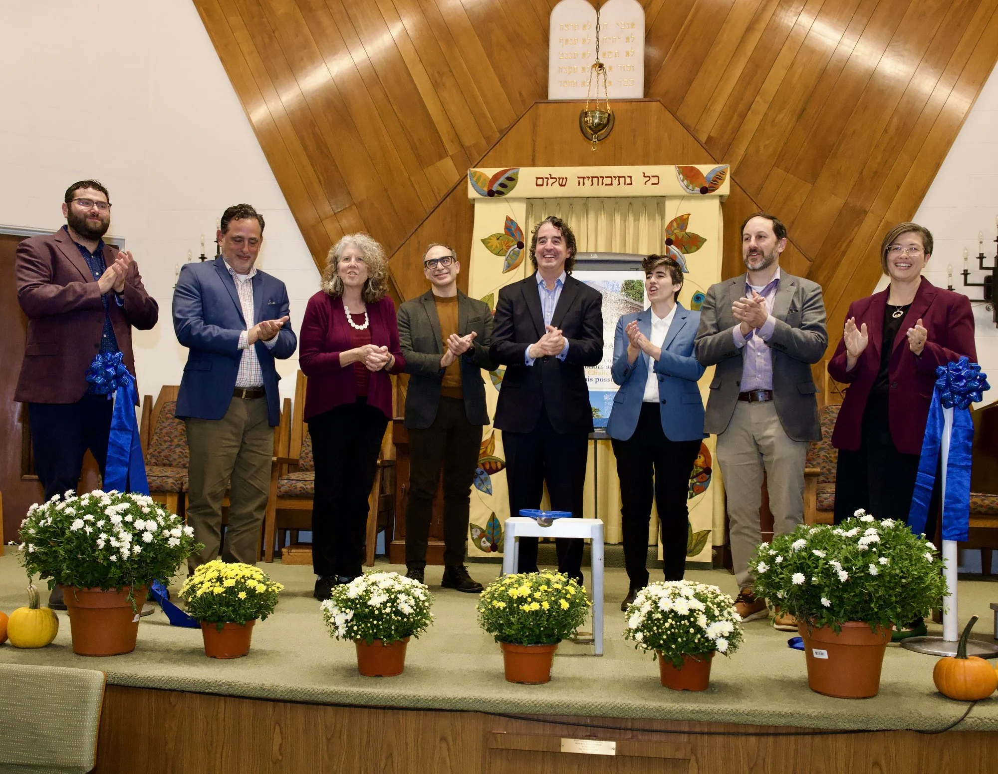 Eight people stand in a row on the bimah applauding, with the cut blue ribbon hanging from the posts at either end. Pots of white and yellow mums line the front of the stage.