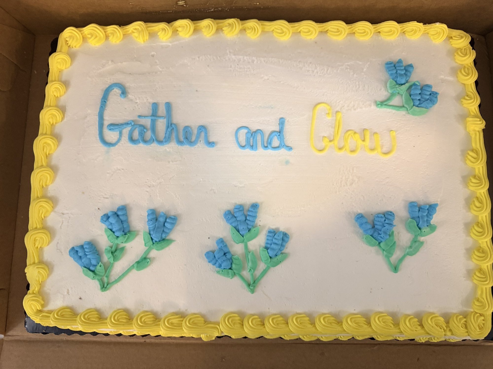 A white sheet cake trimmed in yellow frosting, reading &ldquo;Gather and Glow&rdquo; in blue and yellow icing, decorated with blue flowers.