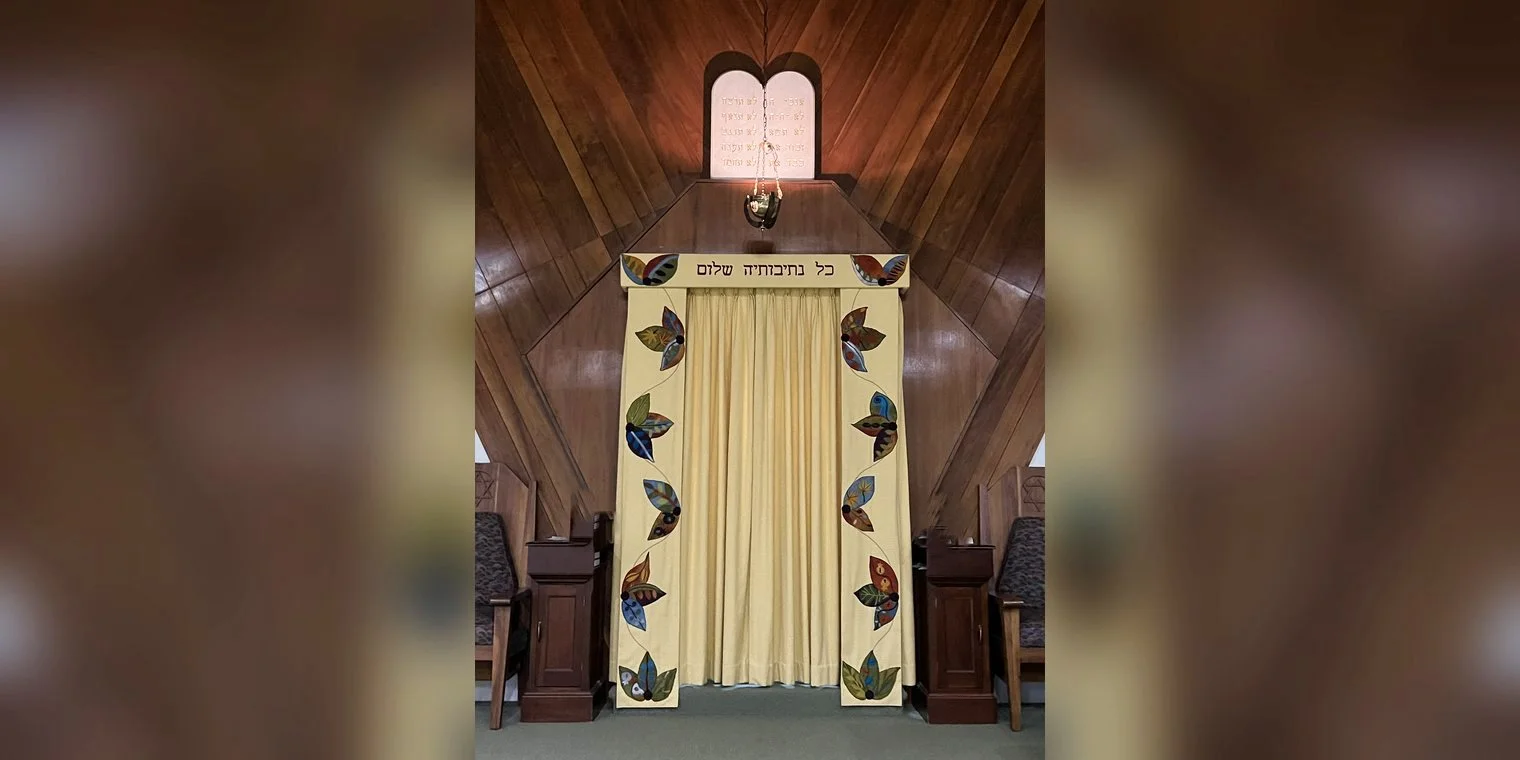 The ark curtain (parochet) in the Temple HBT sanctuary: a golden curtain framed by two panels of colorful leaf appliqué, with a Hebrew banner across the top, and the Ten Commandments tablets and the hanging eternal light above it.
