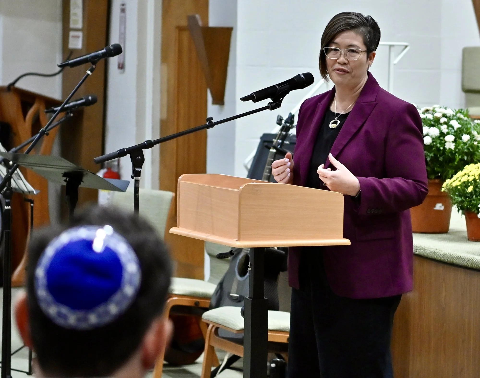 Persis Yu, in a plum blazer and glasses, speaks warmly at the lectern, gesturing with both hands.