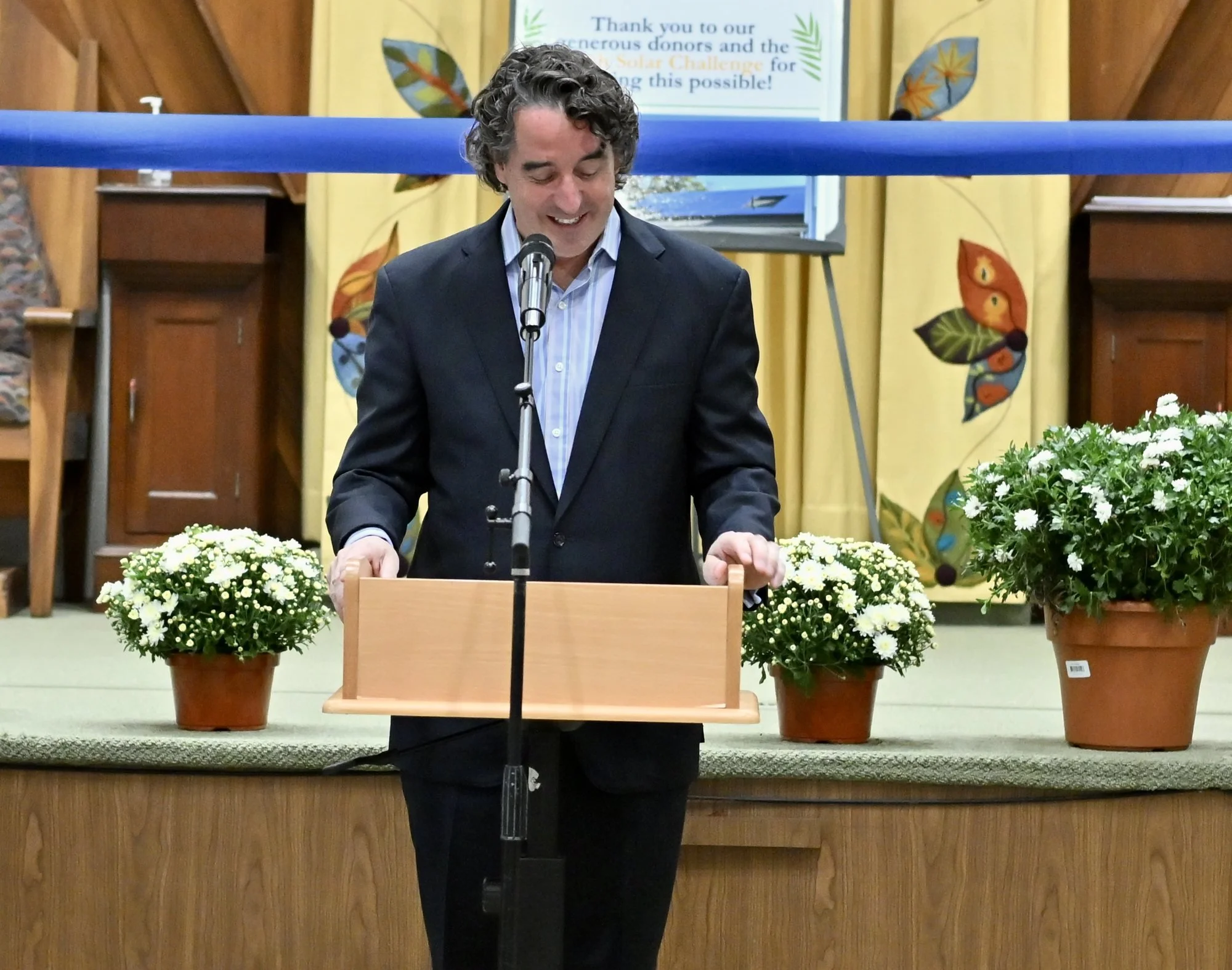 Jason Arenburg, in a dark suit, smiles as he speaks at the podium between pots of white mums.