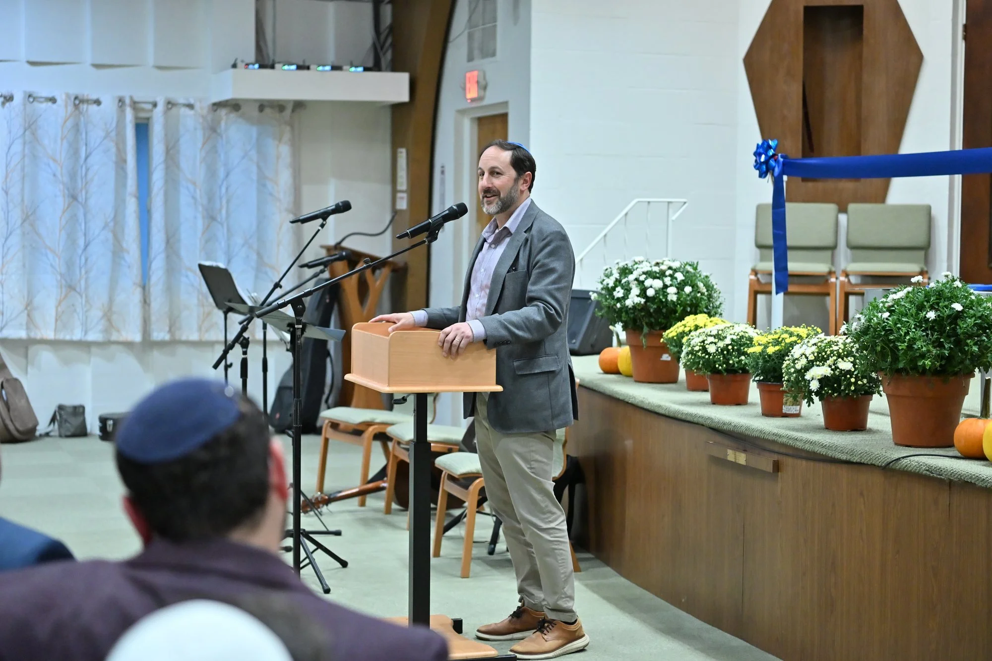 Councilor Benjamin Weber, in a gray jacket, speaks at the lectern beside a row of potted mums.