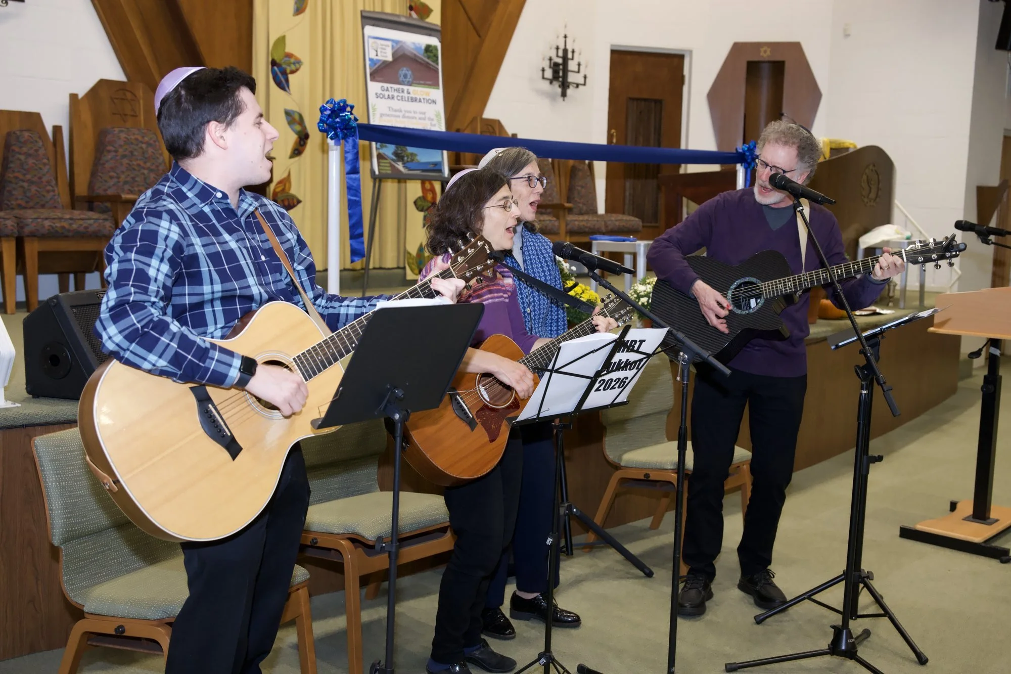 The HBT band sings at microphones in front of the bimah, three of them playing acoustic guitars.