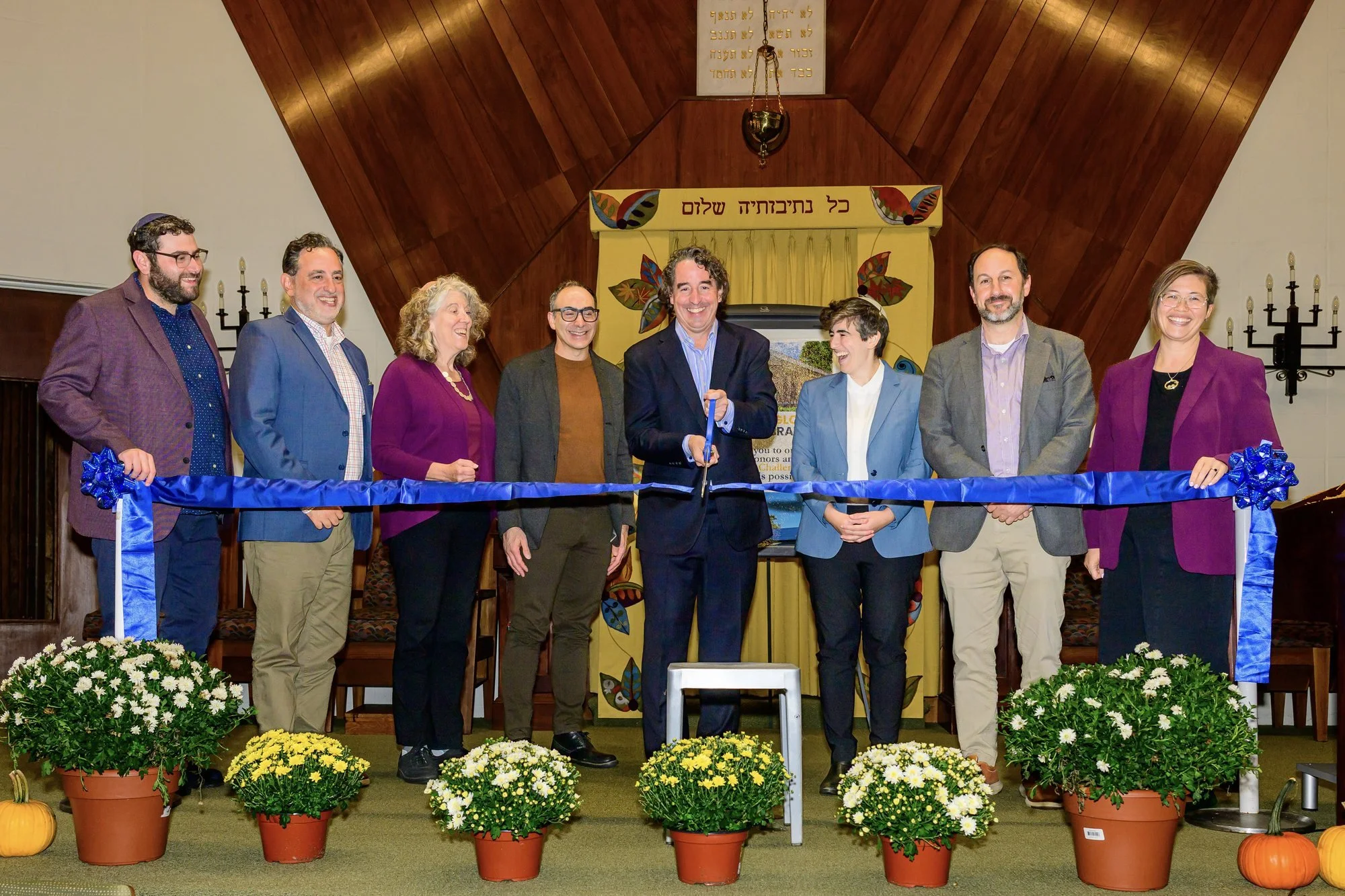Jason Arenburg, center, cuts the blue ribbon with large scissors while seven speakers and clergy stand beside him, smiling.