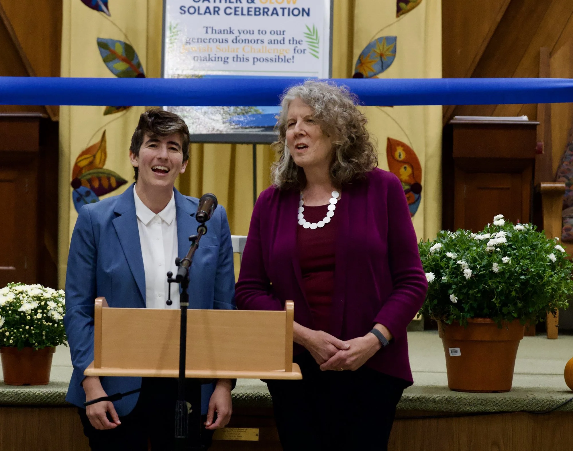 Rabbi Aliza Schwartz, in a blue blazer, and Rabbi Emerita Barbara Penzner, in a plum jacket, stand side by side at the podium, smiling as they sing.