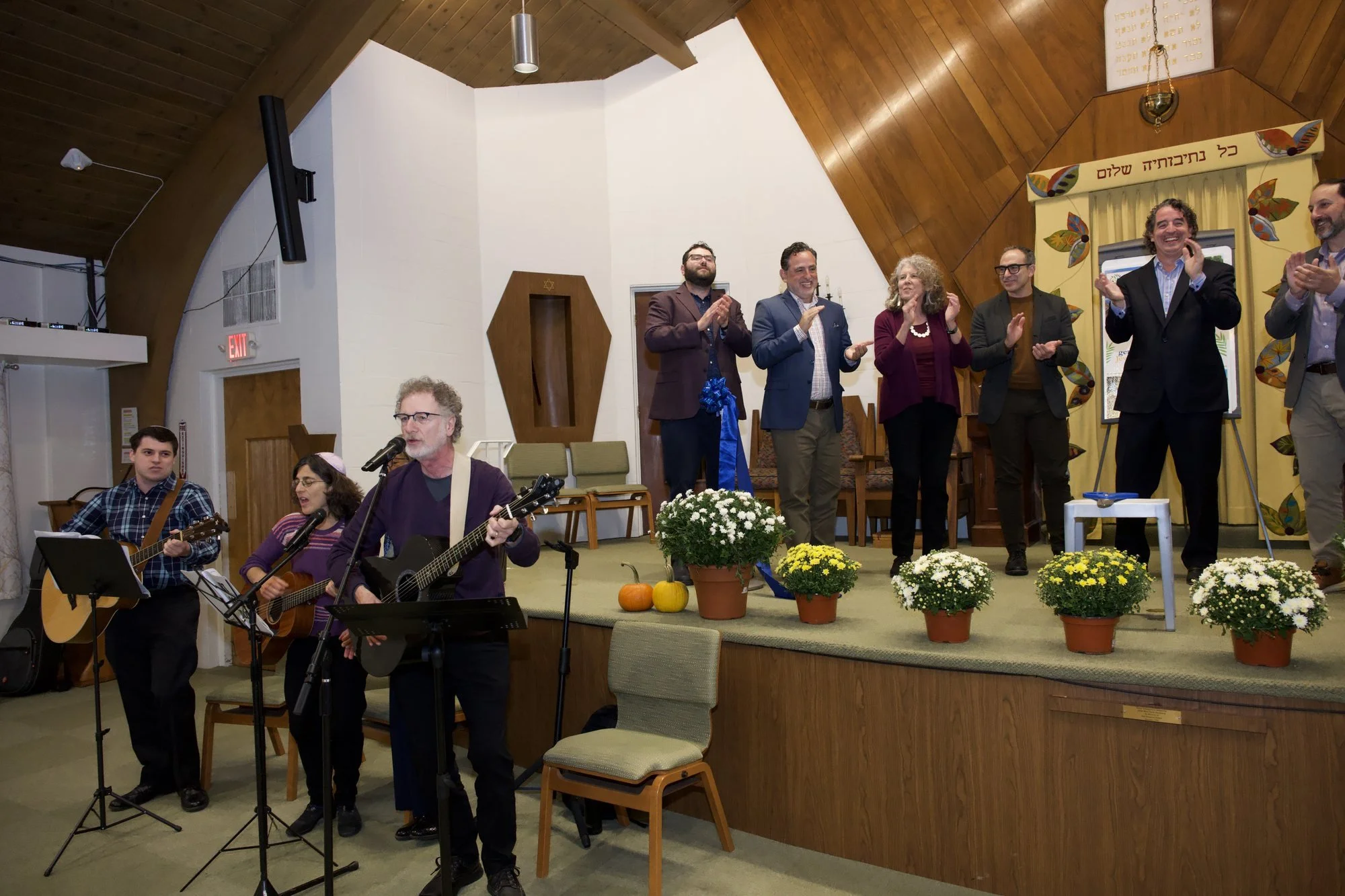 The HBT band plays and sings in front of the bimah while the speakers applaud on the stage behind them.
