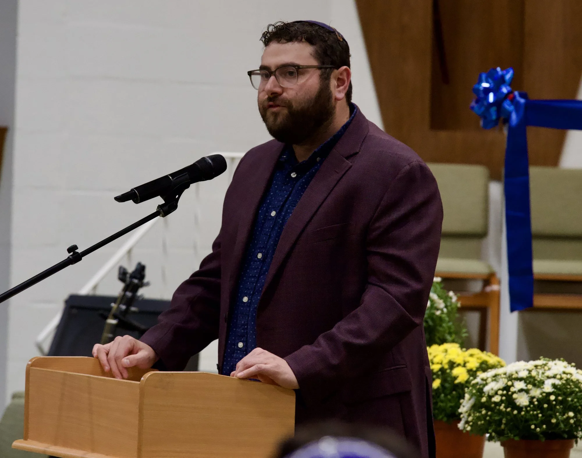 Noah Weinberg, bearded and wearing a kippah and a maroon jacket, speaks at the podium.
