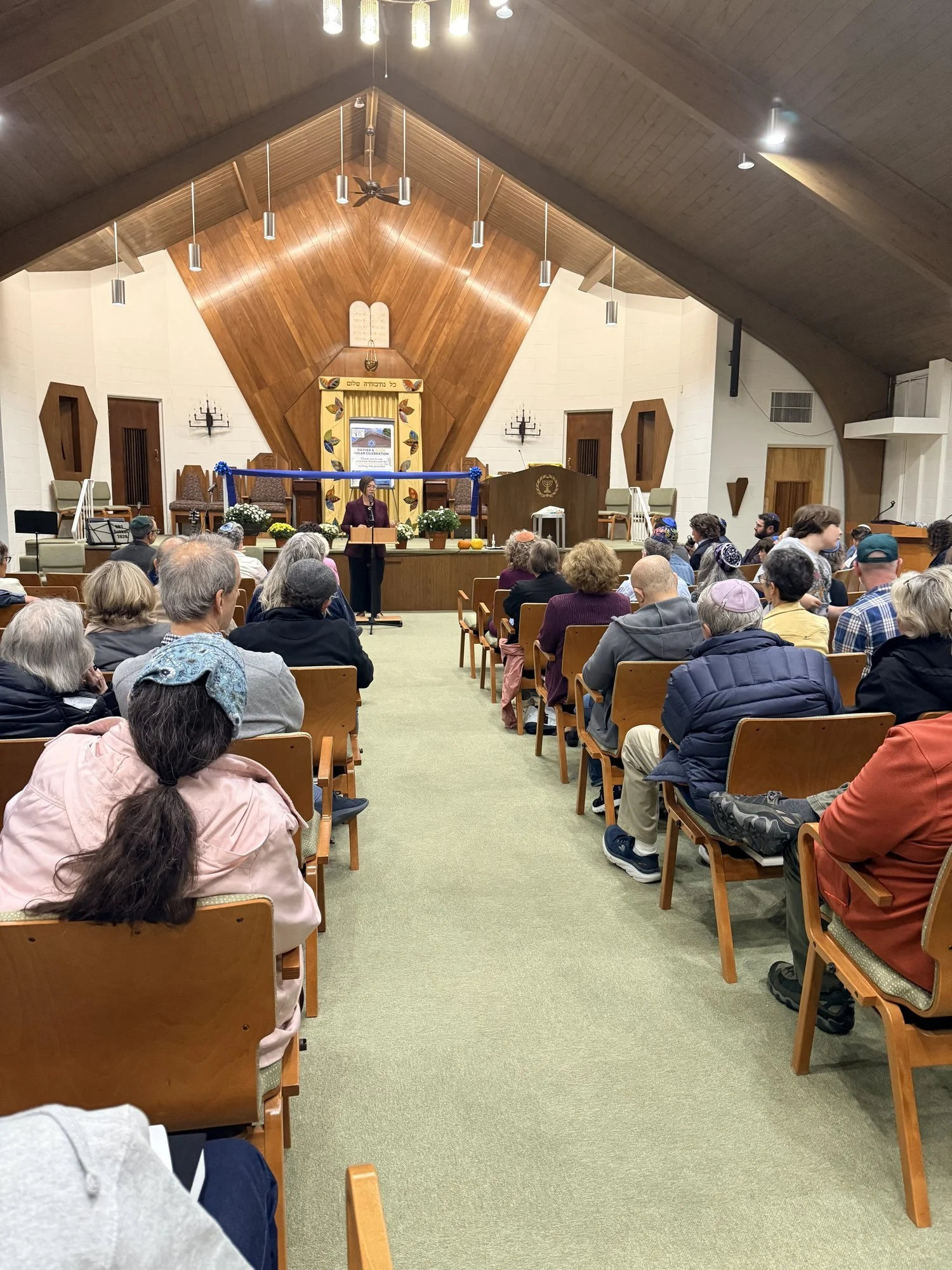 A packed sanctuary seen from the back: rows of people on both sides of the center aisle face the bimah, where a speaker stands at the lectern.