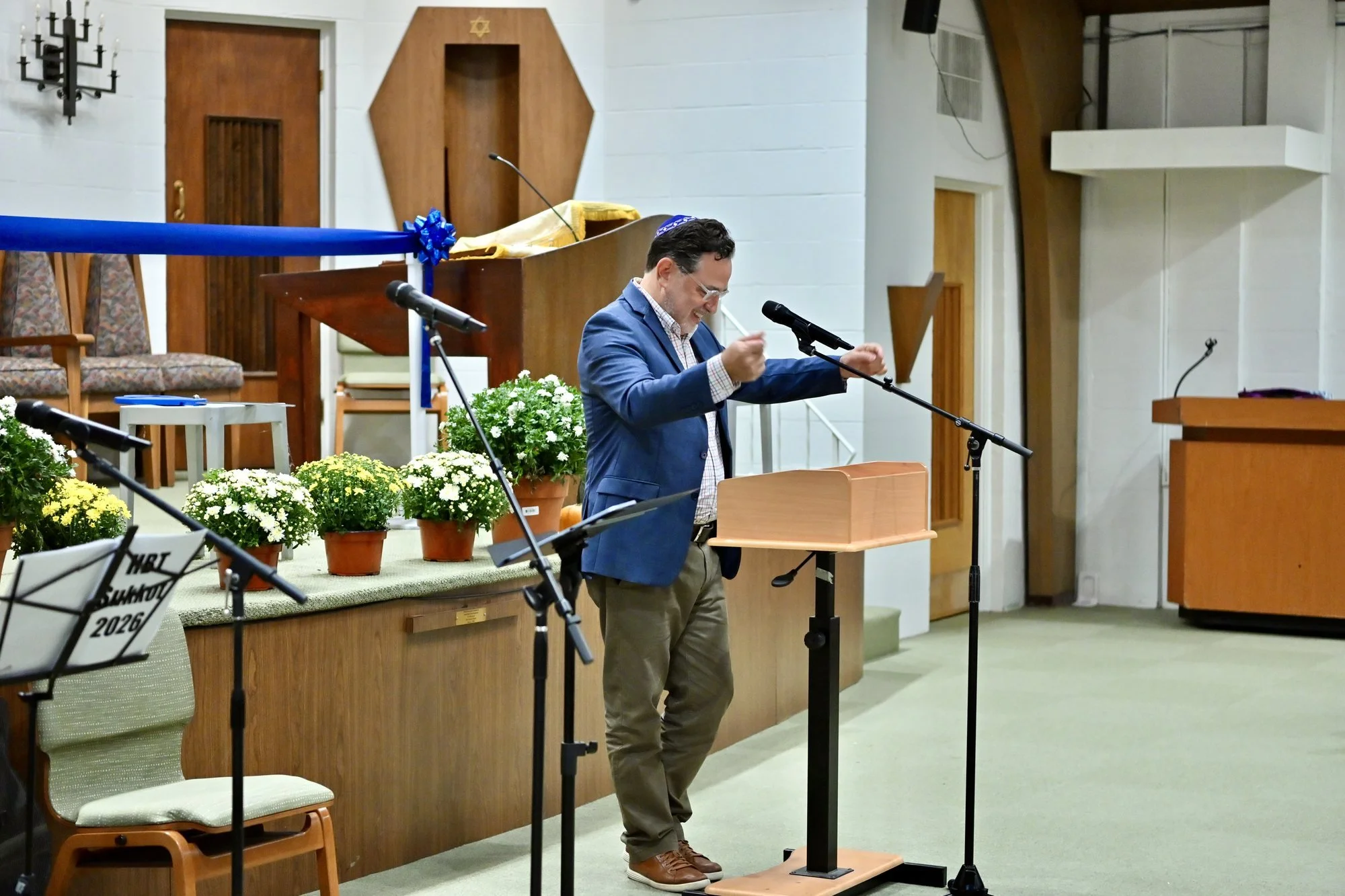 Michael Kadish, in a blue blazer, leans toward the microphone and gestures as he speaks at the lectern beside the bimah.