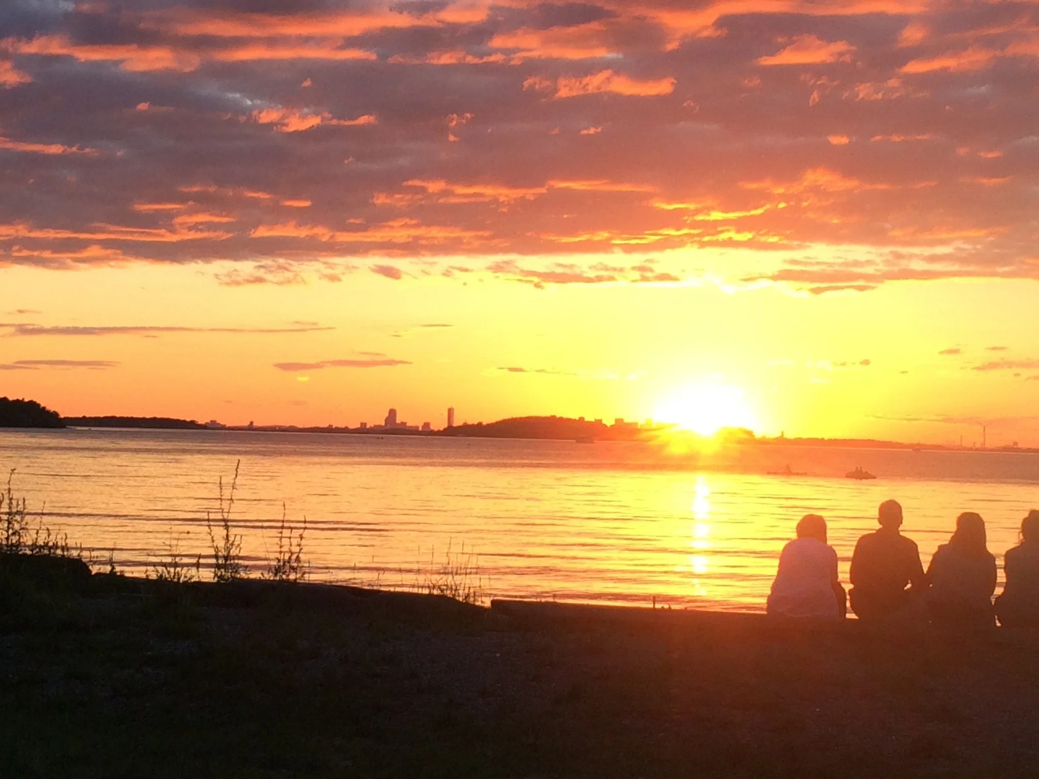 Four people sit side by side on the shore, seen from behind, watching the sun set over Boston harbor. The sky glows orange and pink above the water, with the city skyline small in the distance.