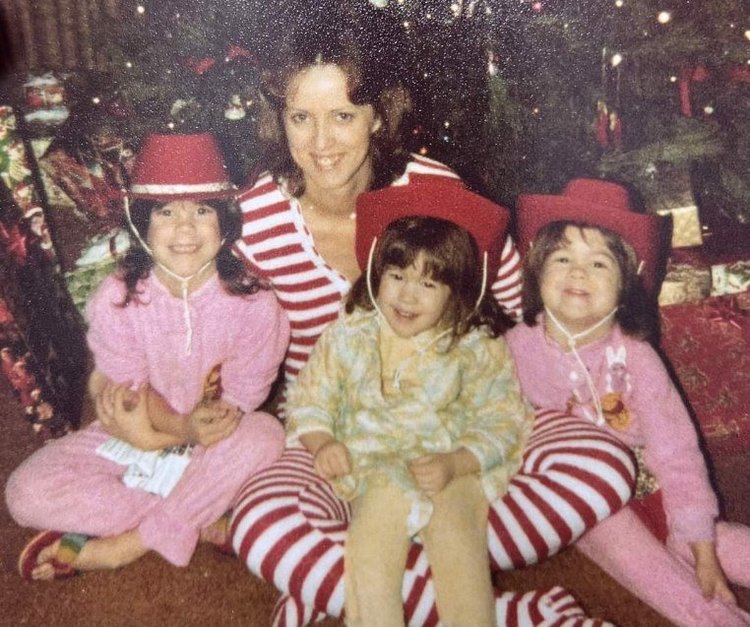 Three young girls in pajamas and cowboy hats, sitting around their mother who is in candy cane pajamas, in front of a Christmas tree
