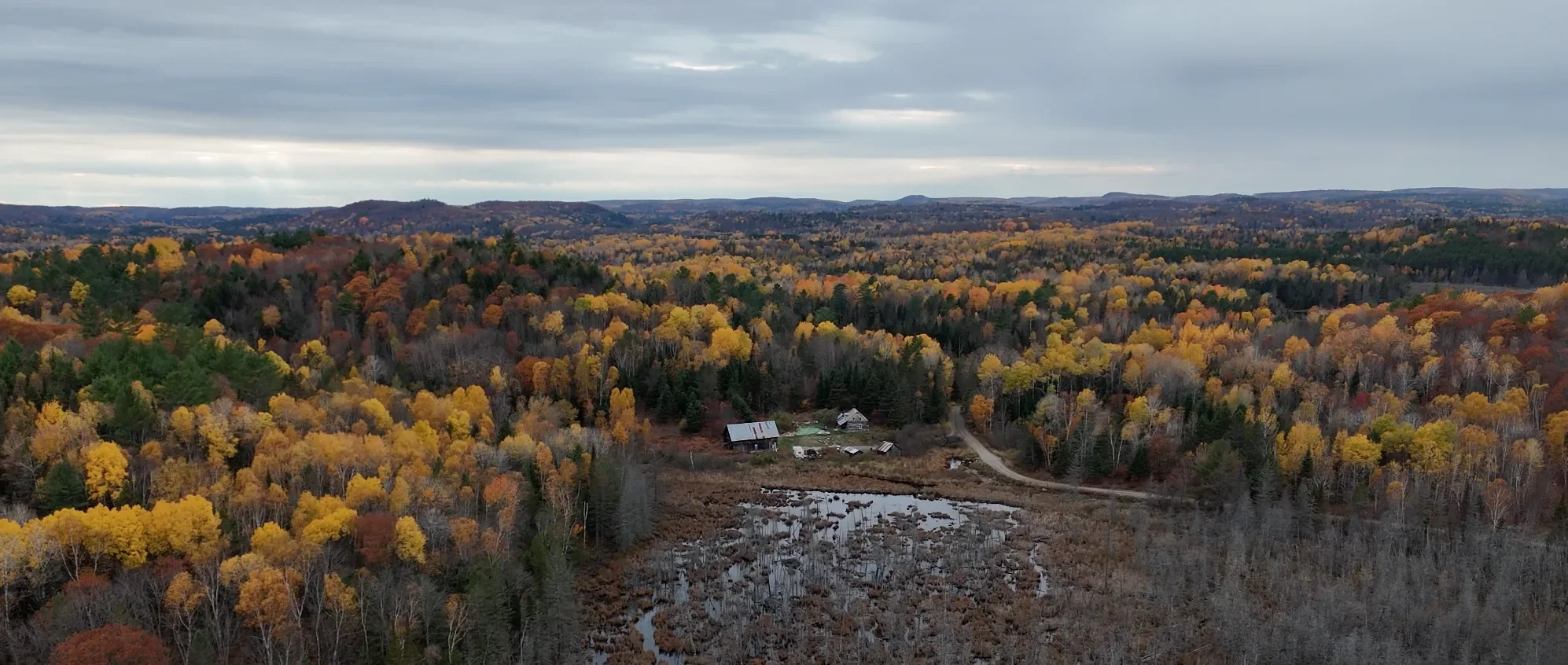Vue a&eacute;rienne de la propri&eacute;t&eacute; LeTerrain, quelques petits toits dans une clairi&egrave;re, entour&eacute;s de tous c&ocirc;t&eacute;s par la for&ecirc;t de feuillus d&rsquo;automne jusqu&rsquo;aux collines lointaines