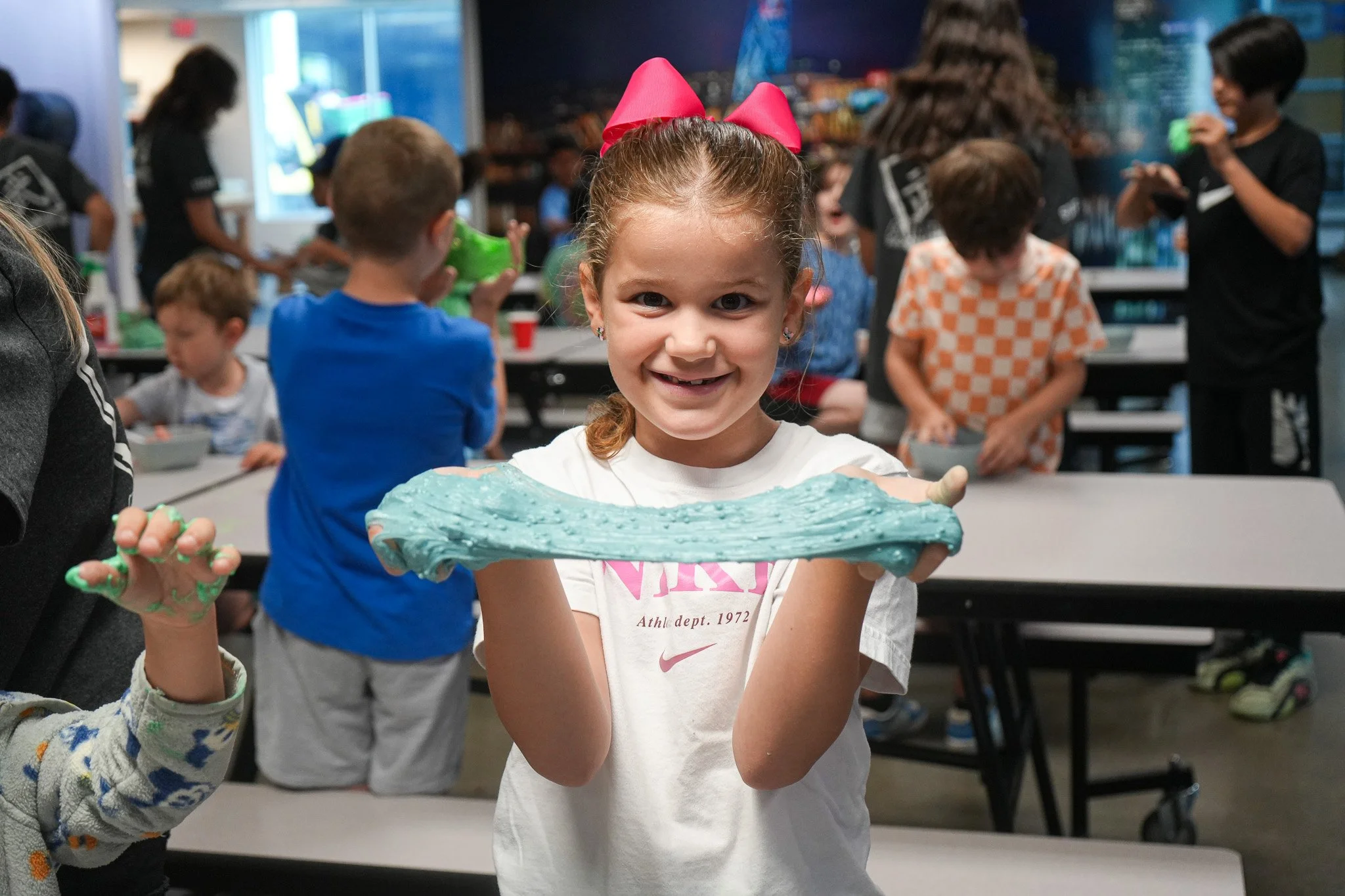 A girl with a pink hair bow smiles broadly while showing how her handmade blue slime stretches.