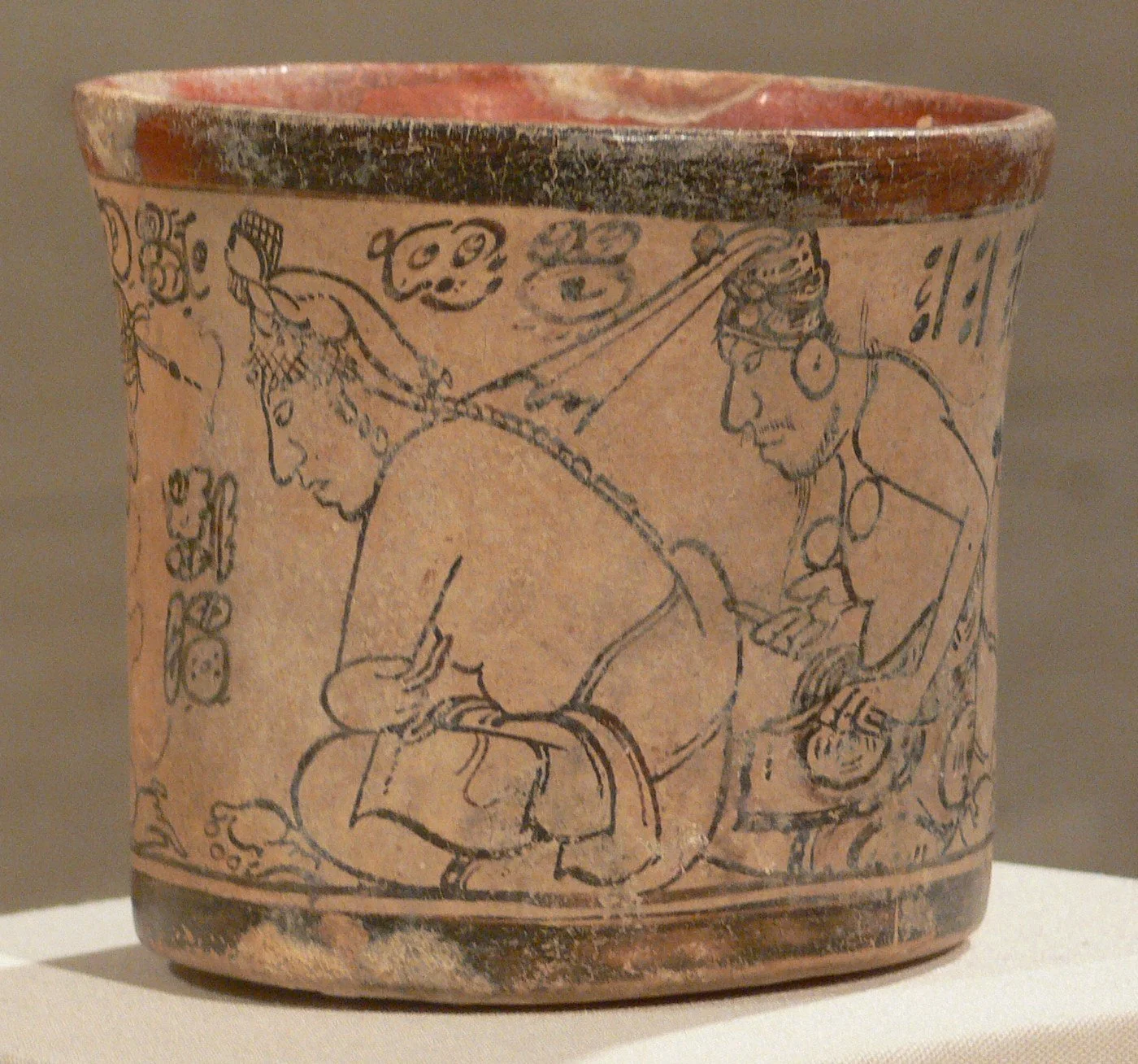 Vaso maya de estilo códice, en negro sobre crema: un joven escriba sentado con un tocado de flores y, detrás, un anciano con tocado de red que se inclina hacia él
