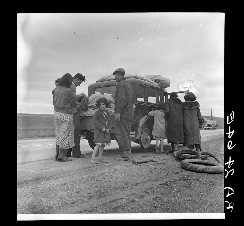 A family stands beside a car loaded with belongings on a roadside