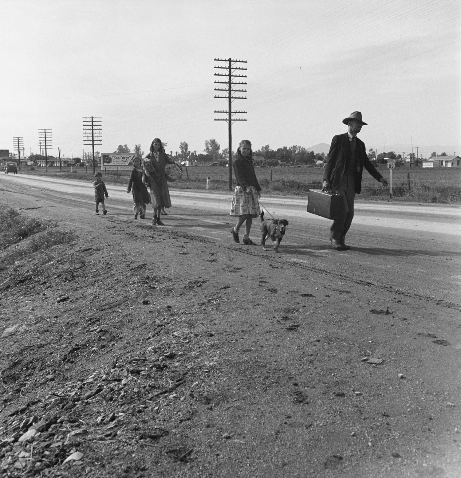 A mother, father and children walk along the shoulder of a highway carrying bundles
