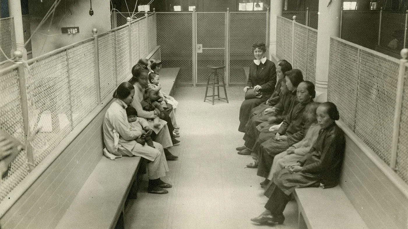 Detainees sit on benches along a fenced corridor