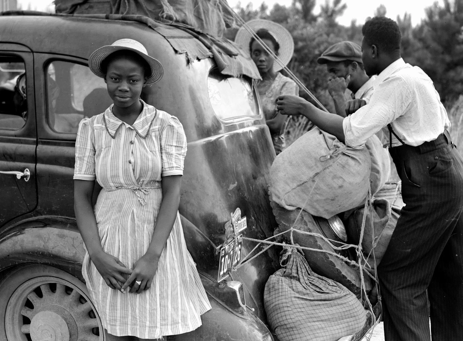 A young woman stands beside a car as a man ties bundles to it