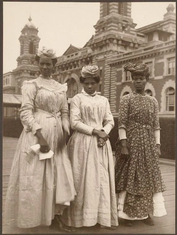 Three women in white dresses stand in front of the Ellis Island building