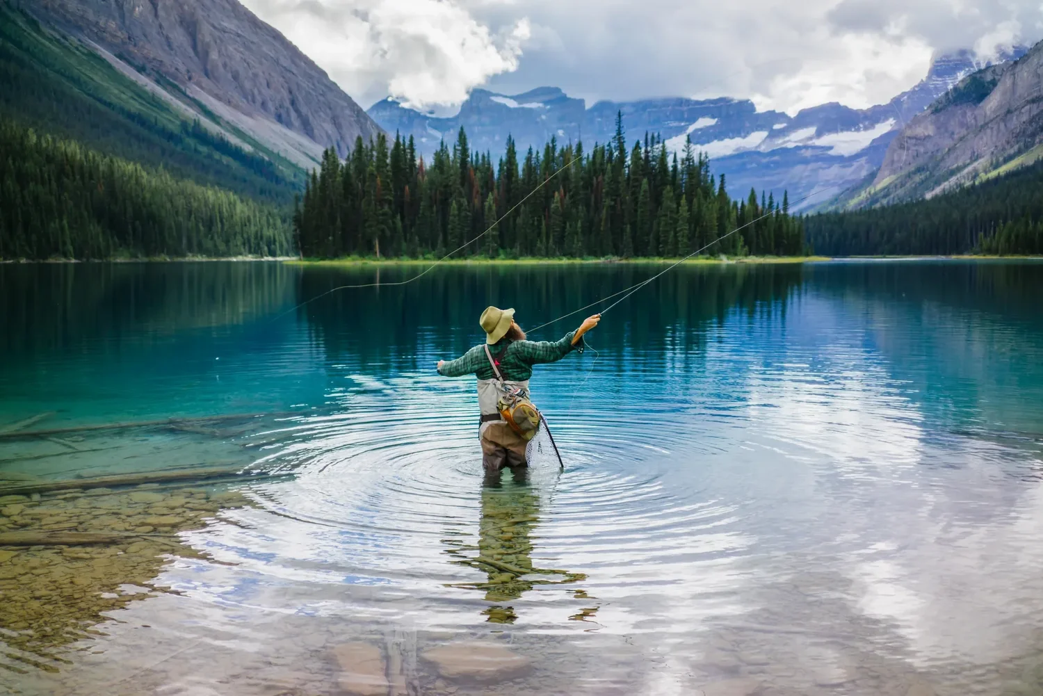 A fly fisherman casting into a still turquoise lake in the Canadian Rockies
