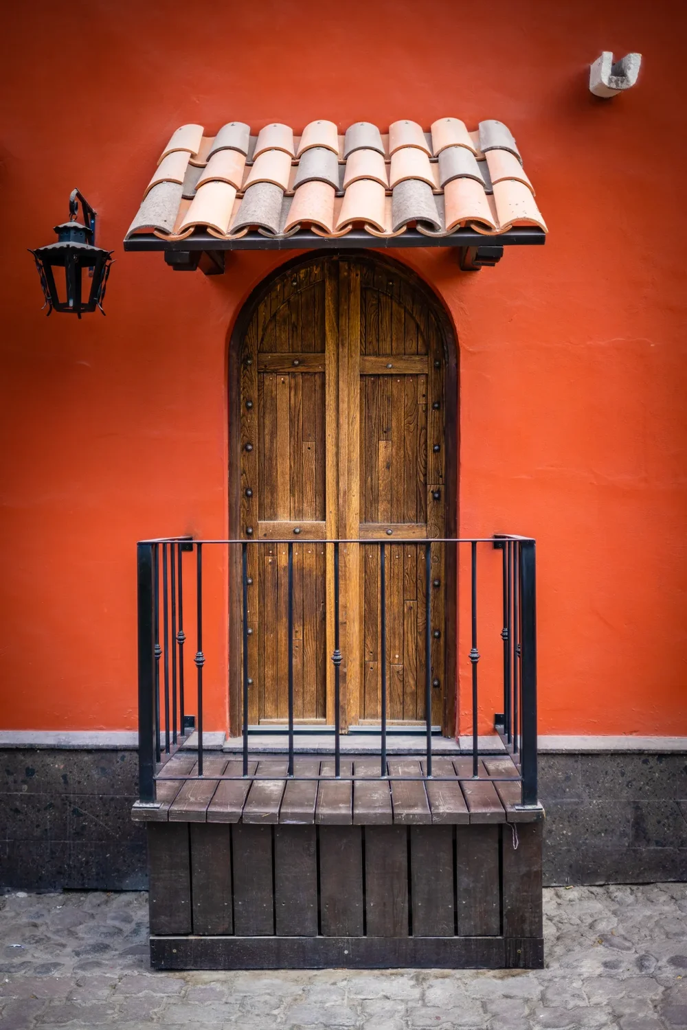 An arched wooden door under a tile awning on a vermillion wall, iron lantern alongside