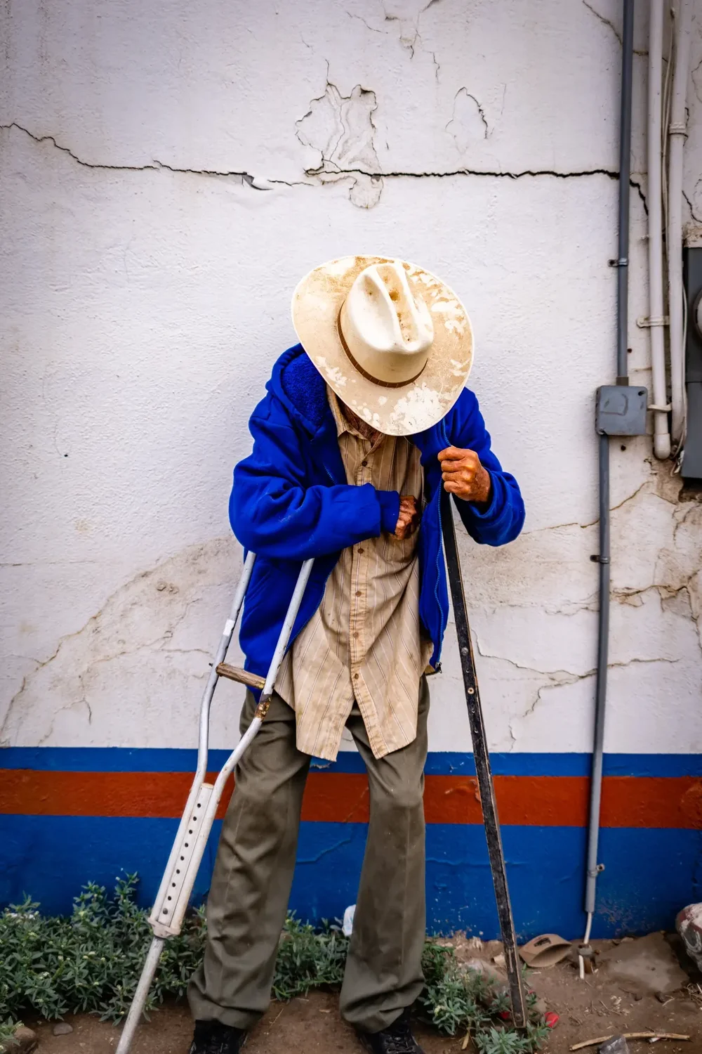 An older man in a straw hat on crutches against a painted wall, Baja