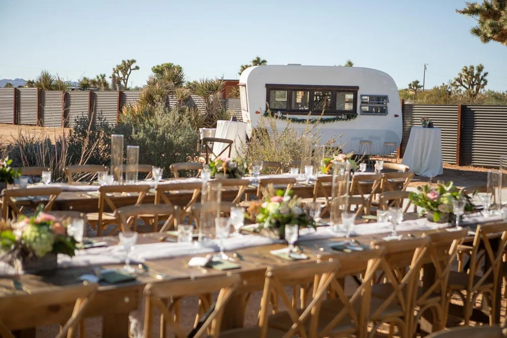 Reception tables with white linens set up in the desert at Serenity Escape, with a vintage trailer and Joshua trees in the background