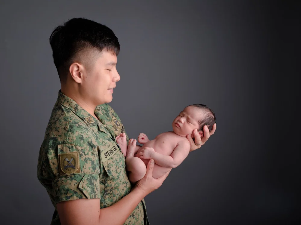 Father in a camouflage uniform gently holding his newborn against a dark grey background, photographed by Mamamiyo Photography in Singapore.