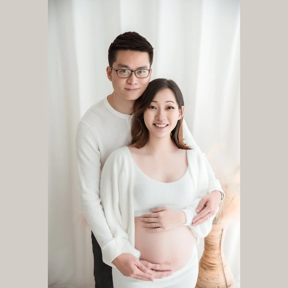 Smiling expectant parents in white, with their hands around the baby bump, photographed by Mamamiyo Photography in Singapore