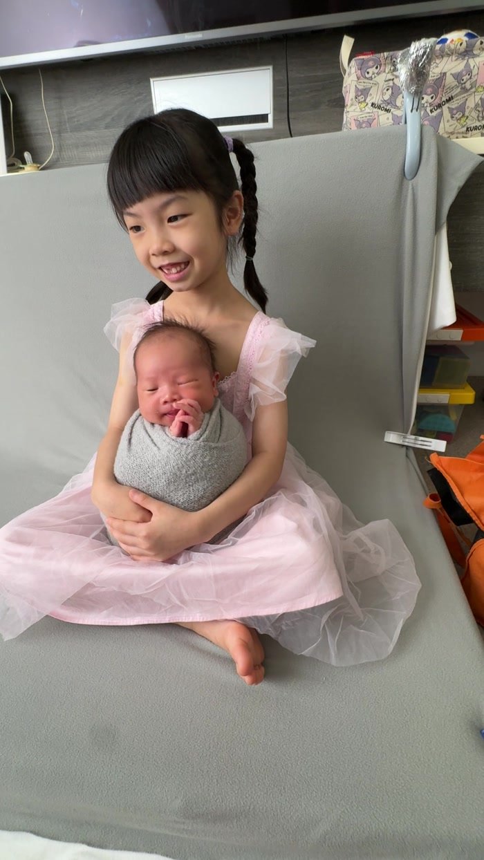 Older sister smiling while holding her wrapped newborn sibling on the grey backdrop during a real Mamamiyo home newborn photoshoot in Singapore.