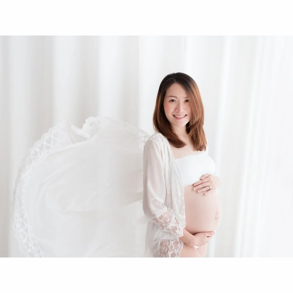 Smiling mother-to-be showing her baby bump in a white lace robe beside curtains, photographed by Mamamiyo Photography in Singapore