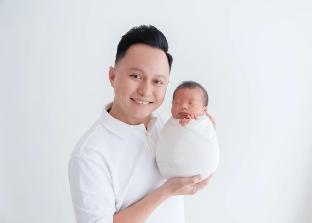 Smiling father in a white shirt holding his sleeping newborn in a white wrap, photographed by Mamamiyo Photography in Singapore.