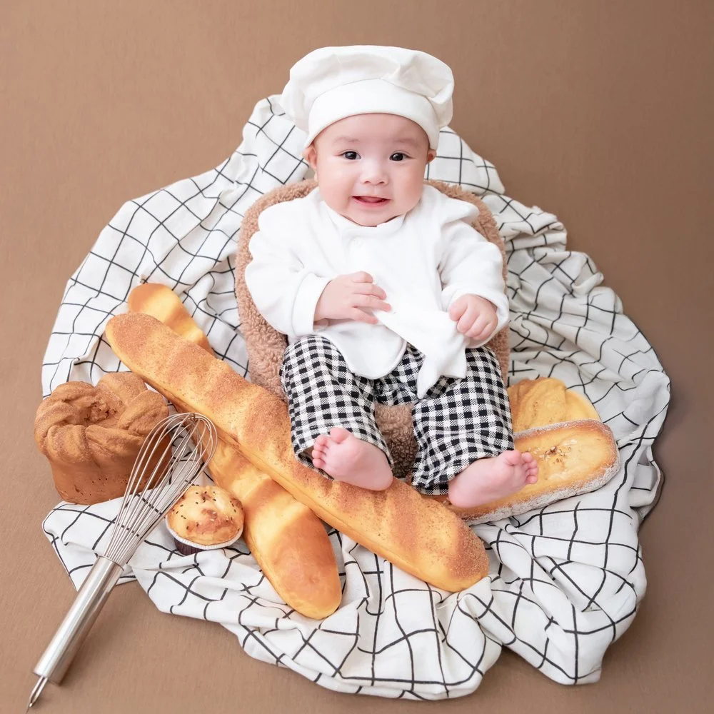 Smiling baby reclining in a chef outfit on a checked blanket beside bread props and a whisk, photographed by Mamamiyo Photography in Singapore.