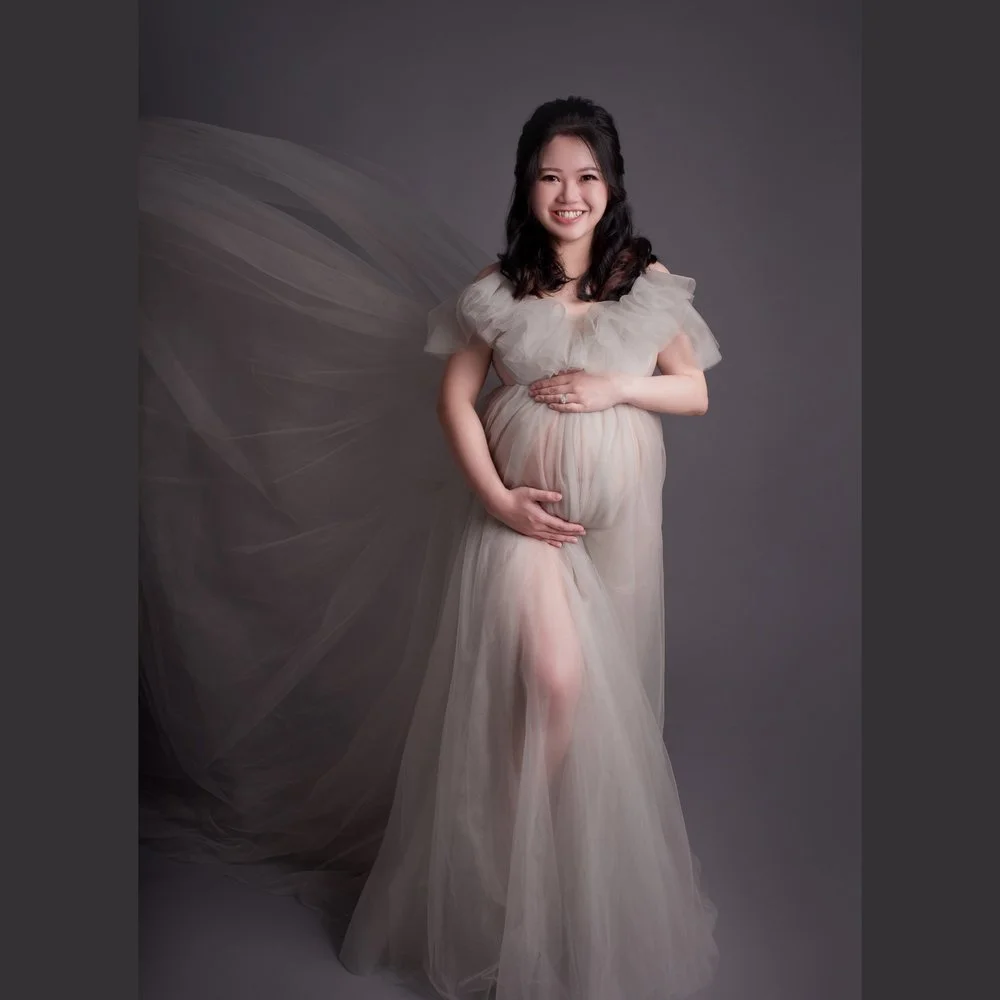Smiling pregnant woman cradling her bump in a grey tulle gown with flowing fabric, photographed by Mamamiyo Photography in Singapore